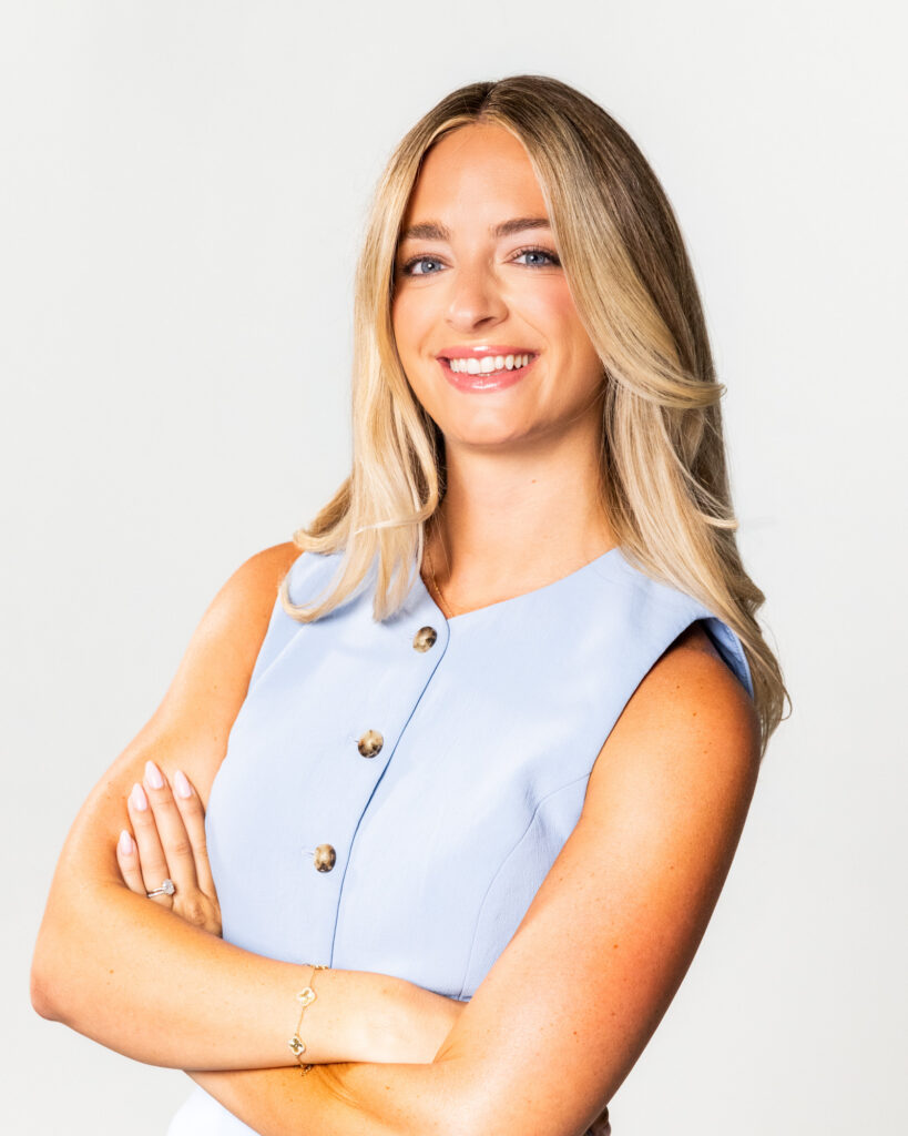 Smiling woman with blonde hair wearing a light blue sleeveless top, arms crossed against a pale background.