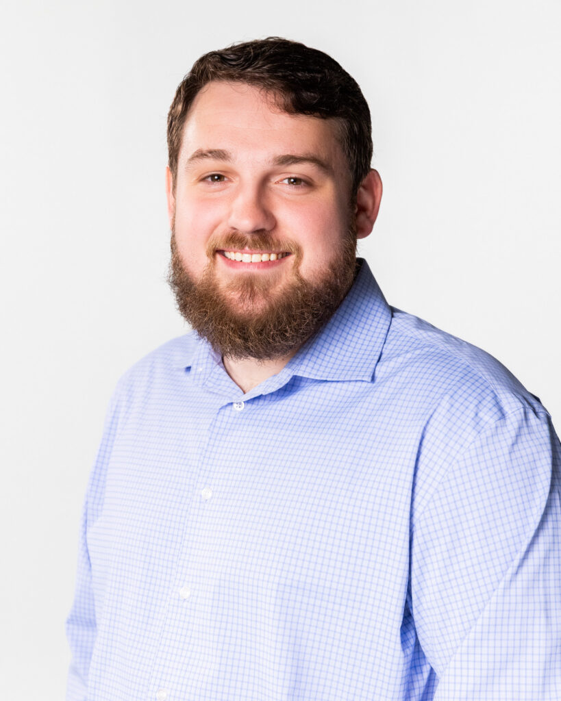 Portrait of a smiling man with a beard wearing a light blue check shirt against a white background, cropped at the shoulders.